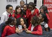 A group of primary school children in red uniform surrounding a female teacher who is holding a mobile phone